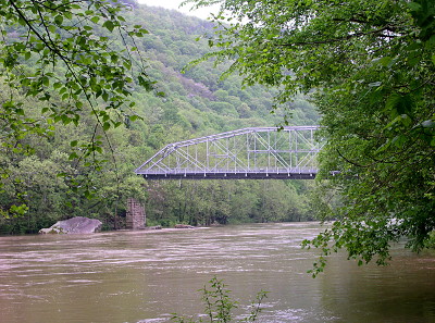 [This image was taken from a river bank just downstream from the bridge. A leafy tree blocks the right half of the bridge. The bridge has what appears to be a brick support at the river's bank and an arch support structure above the road. The water level is high and dirty.]